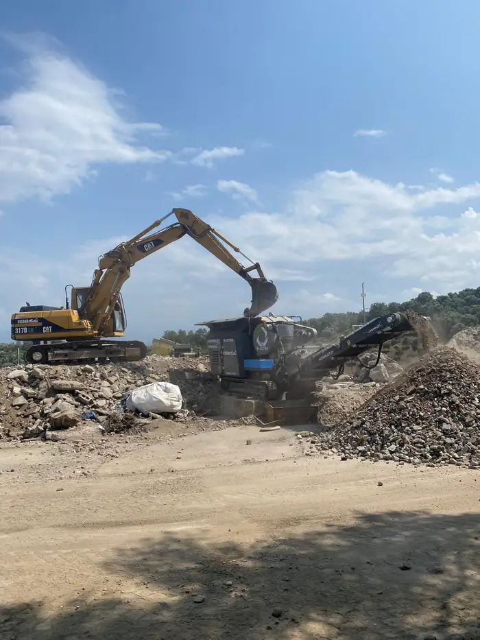 Escavatore cingolato che carica la tramoggia del frantoio mobile mentre il nastro trasportatore scarica il materiale lavorato