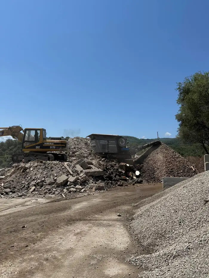 Frantoio mobile in funzione nell'impianto, con cumulo di macerie da demolizione a sinistra e cumulo di aggregato lavorato a destra