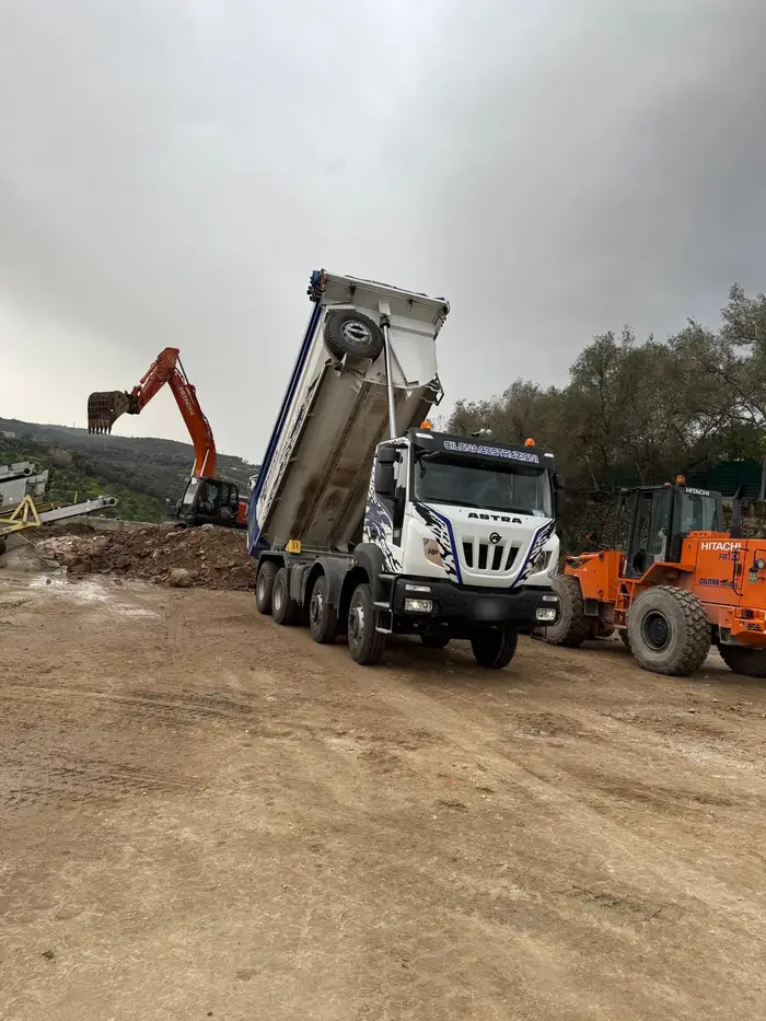 Autocarro a quattro assi con cassone ribaltabile sollevato durante lo scarico, affiancato da pala gommata ed escavatore