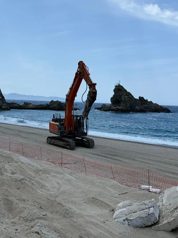 Escavatore cingolato con martello demolitore al lavoro sull’arenile davanti al mare, con scogli affioranti sullo sfondo e recinzione di cantiere arancione