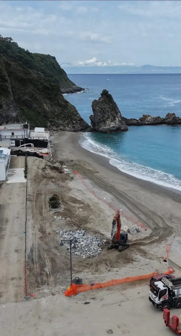 Veduta dall’alto di un cantiere sul litorale, con escavatore, blocchi di scogliera e autocarro dietro la recinzione arancione, affacciato sulla spiaggia