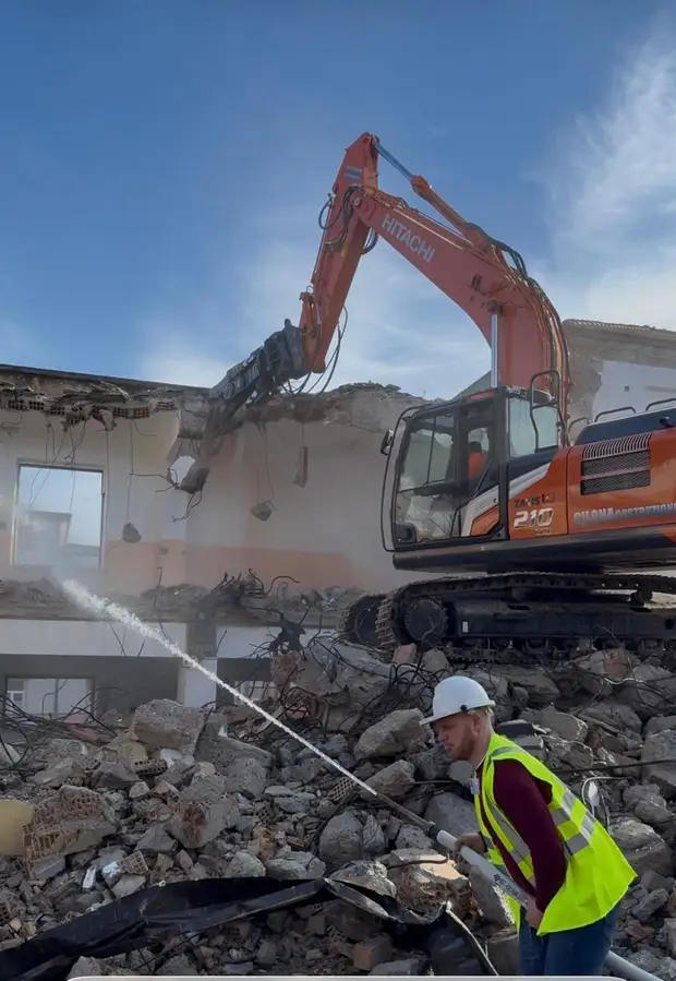 Escavatore cingolato con martello demolitore che abbatte il solaio di una palazzina mentre un operaio con casco e gilet ad alta visibilità bagna le macerie con una lancia d’acqua per contenere la polvere
