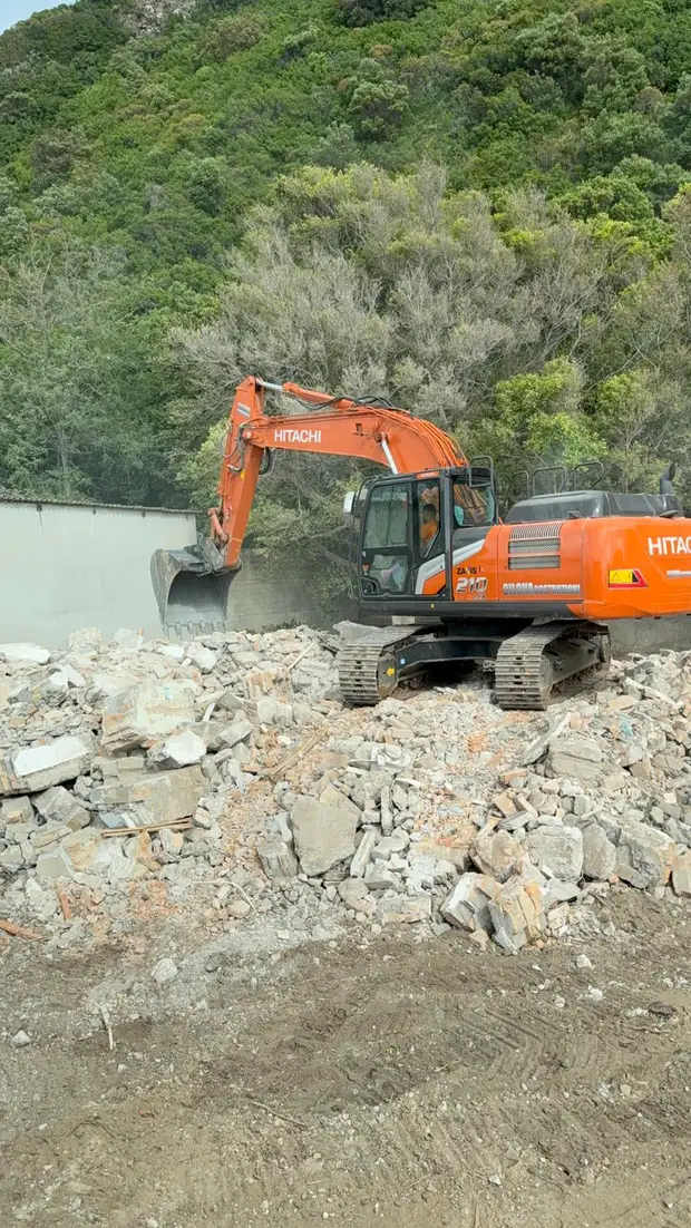 Escavatore sopra un cumulo di macerie di calcestruzzo e laterizio al termine di una demolizione, con un versante boscoso alle spalle