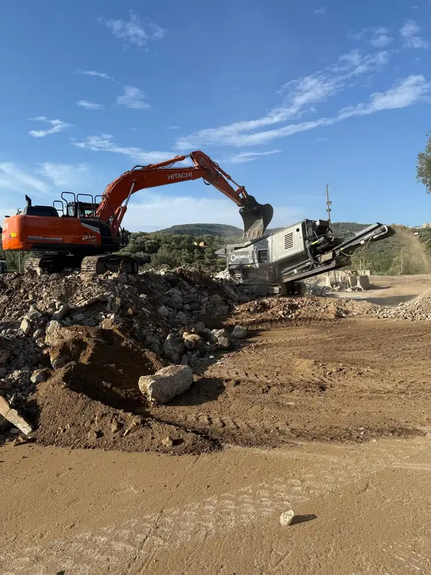Escavatore che alimenta un frantoio mobile in cantiere, accanto a un cumulo di macerie da demolizione