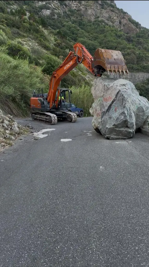 Escavatore cingolato che rimuove un masso di grandi dimensioni caduto sulla carreggiata di una strada di montagna